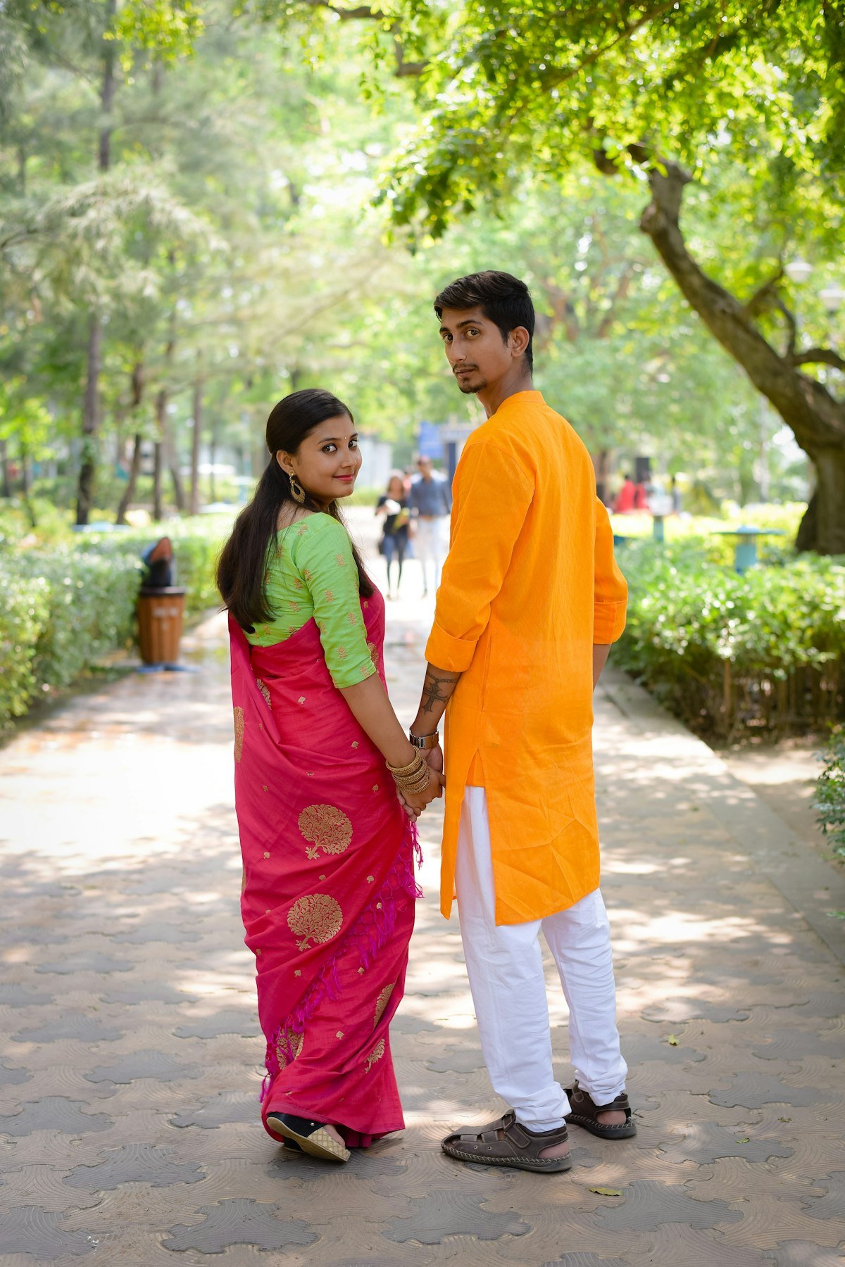 A couple in Indian clothes, the woman in a pink sari and the man in an orange kurta, walking hand in hand on a park path, seen from behind