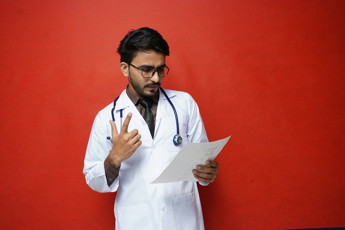 An Indian male doctor in a white coat with a stethoscope reading a paper, against a red background