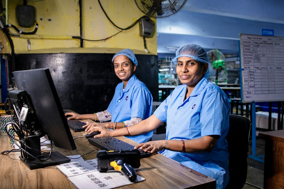Two women in blue work uniforms and caps working on computers in a factory office