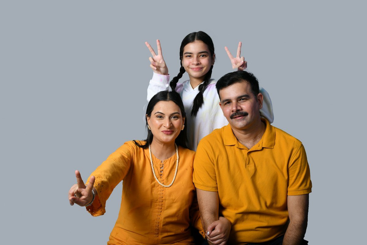 An Indian family of three, a mother, father and teenage daughter in yellow and white, smiling against a grey background