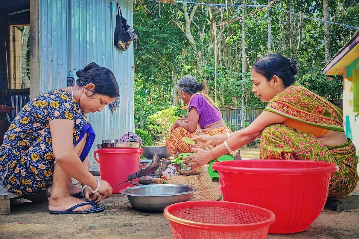 Women preparing food outdoors beside a house with red tubs and steel bowls